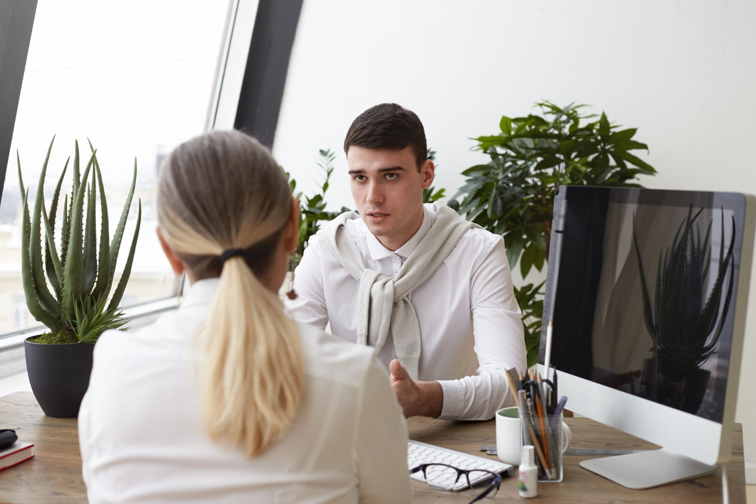 A man and woman sit at a desk in an office setting in professional attire, surrounded by a wall and a houseplant.