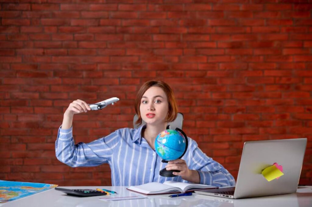 A person in a striped shirt holds a model airplane and a globe at a desk with a laptop and open notebook against a brick wall background.