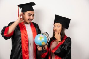 Two graduates in black caps and gowns with red accents hold a globe, expressing excitement and curiosity in a celebratory pose.