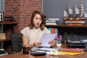 A person sits at a desk, holding papers, surrounded by office supplies, books, and a decorative ship model against a brick wall.