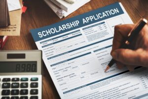 A person fills out a scholarship application form at a wooden desk, accompanied by a calculator and scattered paperwork.