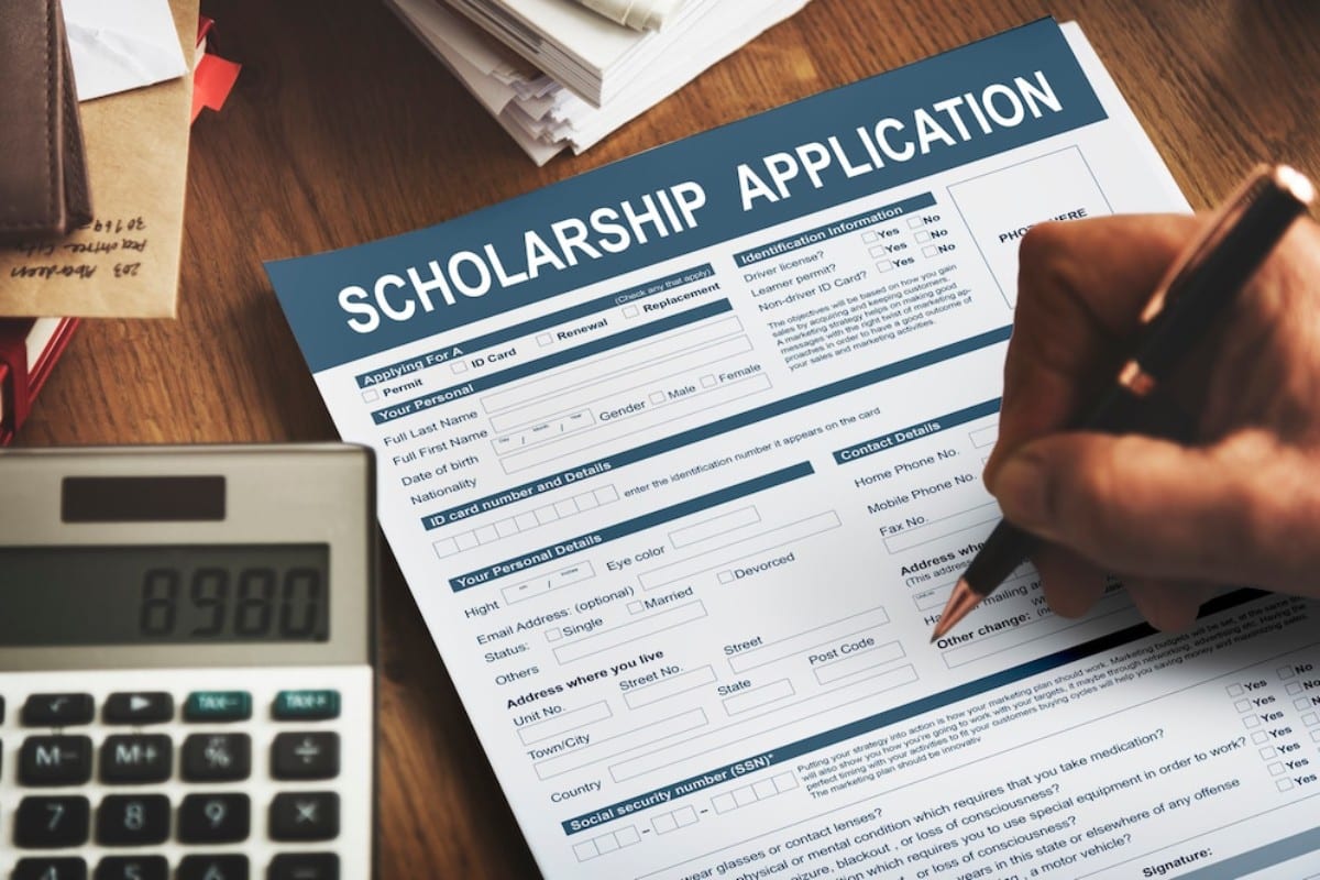 A person fills out a scholarship application form at a wooden desk, accompanied by a calculator and scattered paperwork.