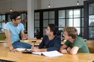 A group of three students engages in a lively discussion at a wooden table filled with books and notes in a modern study space.