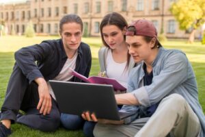 Three people sit on grassy lawn, collaborating on a laptop while one takes notes in a pink notebook, surrounded by greenery and architecture.