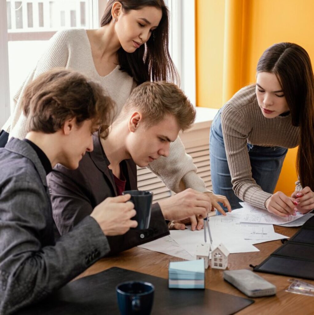 A group of individuals collaborates around a table filled with blueprints and documents, discussing ideas while sipping beverages.