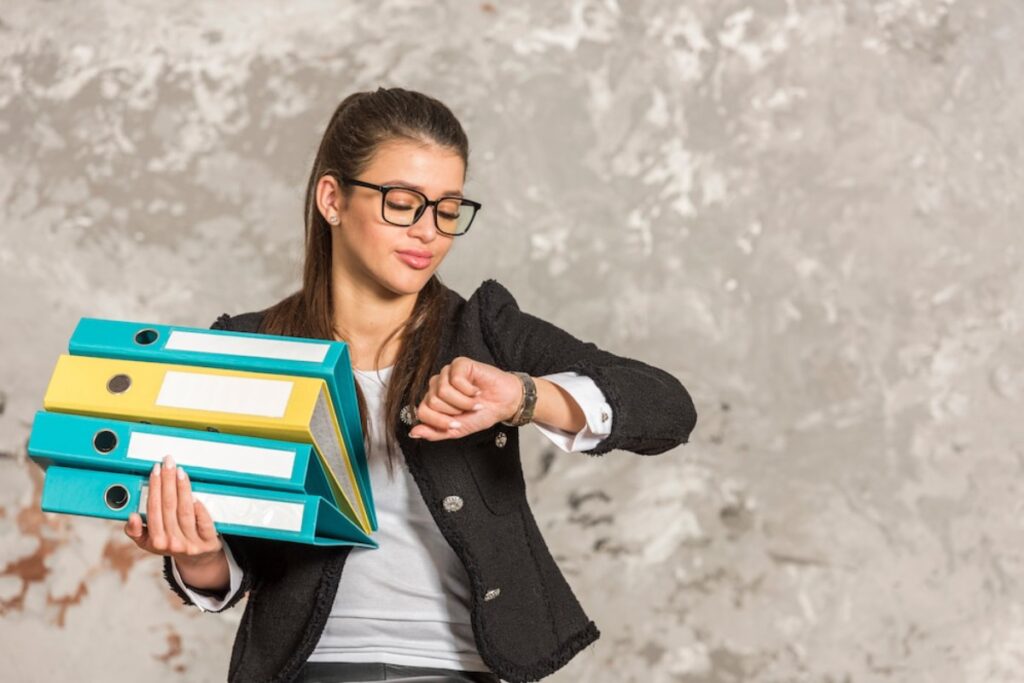 A businesswoman in a black blazer holds three brightly colored binders while checking her watch against a textured wall background.