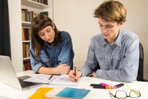 Two students collaborate at a desk with a laptop and study materials, surrounded by bookshelves. Highlighters and notebooks are visible.
