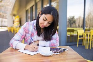 A person writes in a notebook while seated at an outdoor café table with a cup of coffee and a smartphone nearby.