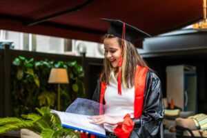 A graduate in a black cap and gown with red accents holds a document, surrounded by greenery and modern decor.