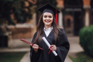 A graduate in a black cap and gown holds a diploma and a certificate, standing in a pathway with greenery and historical architecture behind.
