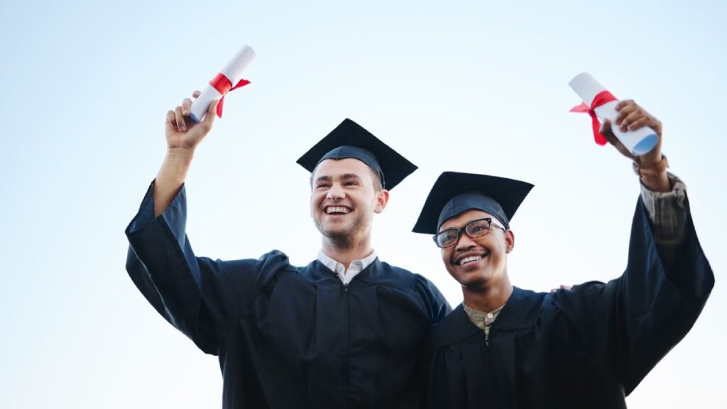 Two graduates in caps and gowns celebrate, holding diplomas with red ribbons against a clear blue sky.