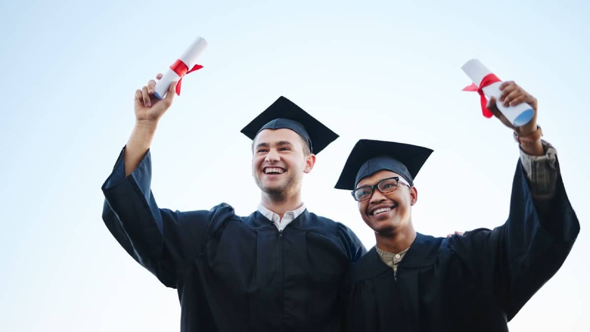 Two graduates in caps and gowns celebrate, holding diplomas with red ribbons against a clear blue sky.
