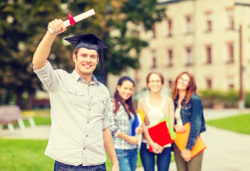 A graduate in a cap holds up a diploma, celebrating with peers holding colorful folders, in a green outdoor setting.
