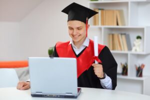 A graduate in a cap and gown sits at a table, holding a diploma and looking at a laptop, with bookshelves in the background.