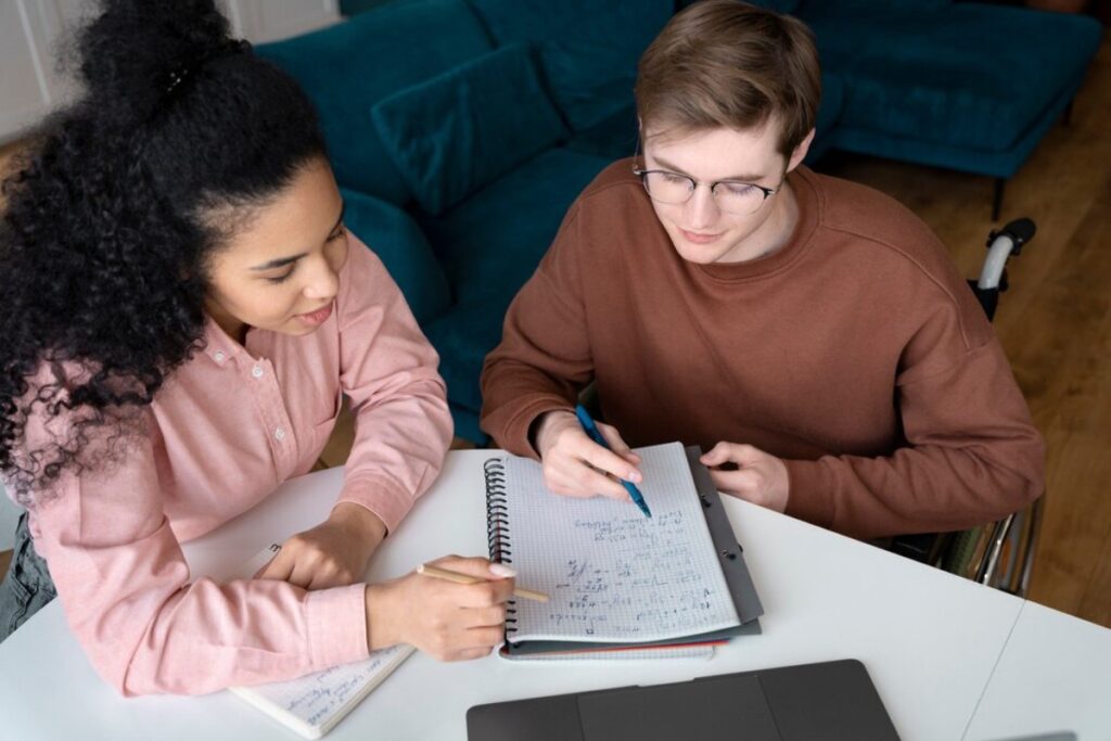 Two individuals collaborate on a study session, writing notes and equations in a notebook, with a laptop on the table and a cozy couch in the background.