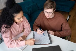 Two individuals collaborate on a study session, writing notes and equations in a notebook, with a laptop on the table and a cozy couch in the background.