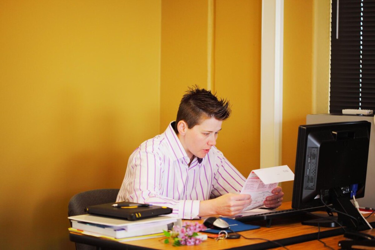 A person in a striped shirt sits at a desk, examining a document, with a computer and books nearby in a warmly colored office.