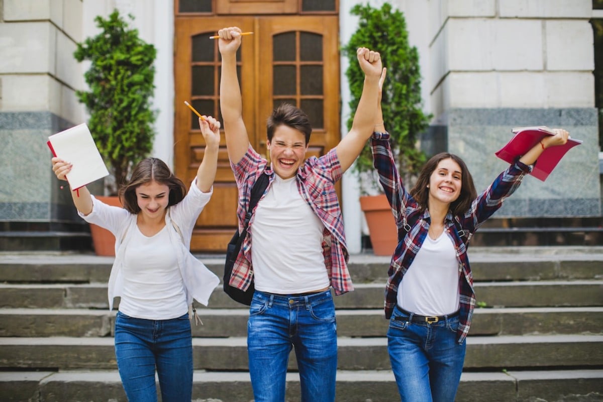 Three excited students celebrate outdoors, raising their arms and holding papers, pencils, and notebooks in front of a building entrance.
