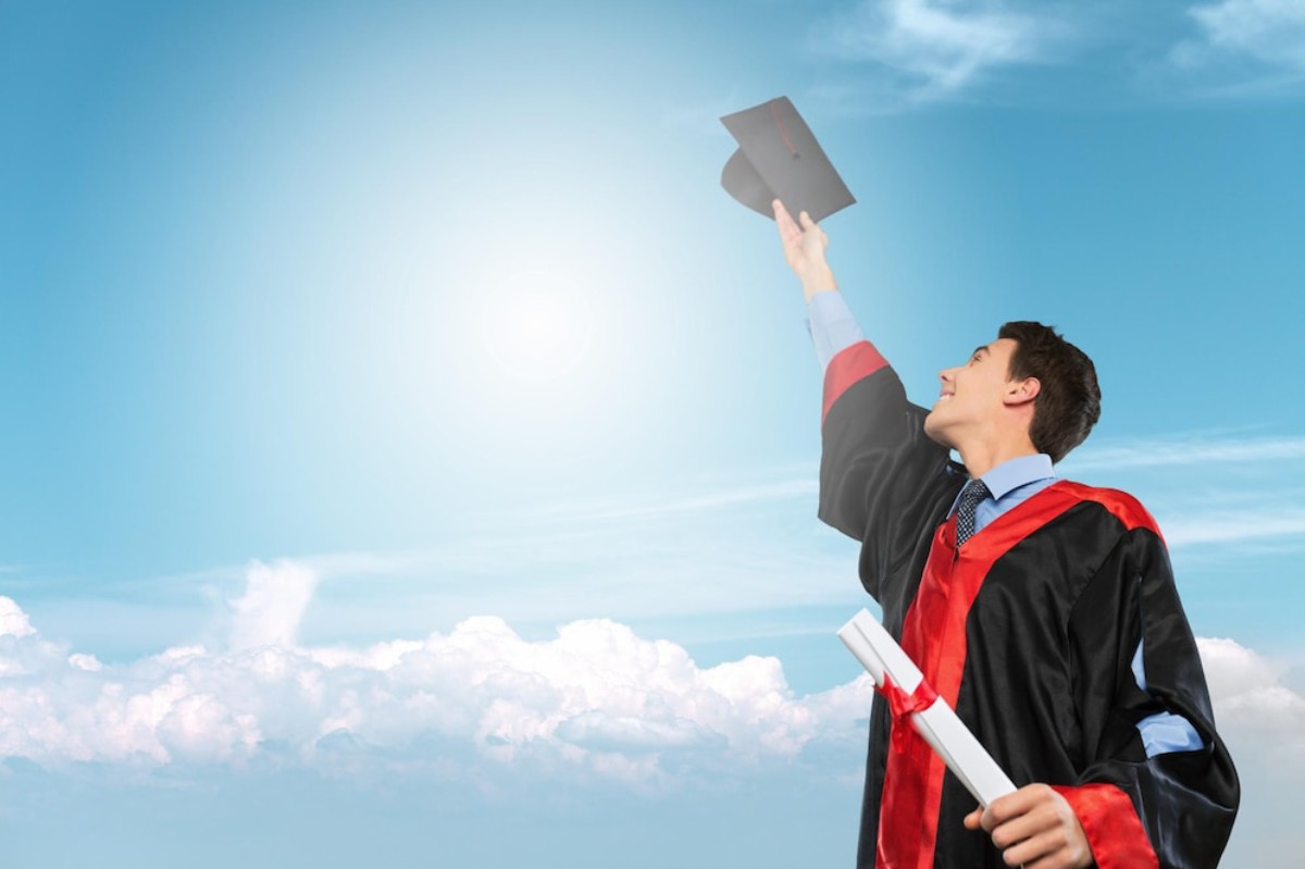 A graduate in a cap and gown joyfully raises a diploma against a bright blue sky with fluffy white clouds.