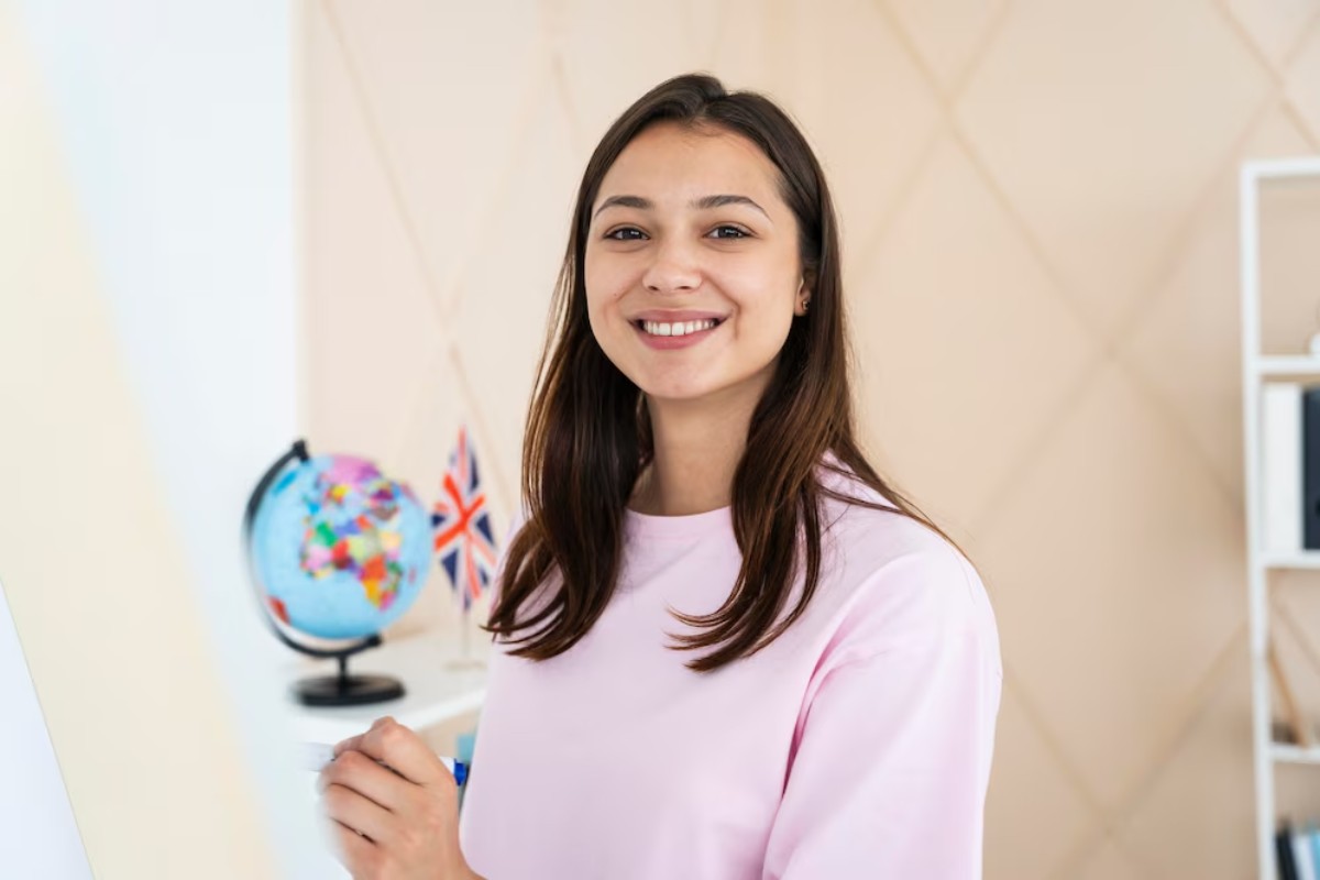 A woman in a pink top stands beside a globe and a British flag, holding a marker in a bright, modern room.