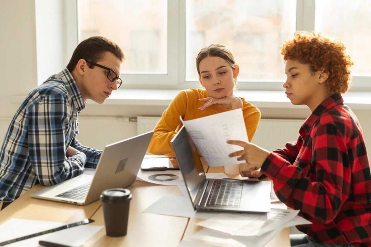 Three people collaborate around a table, reviewing documents and working on laptops, with coffee cups and scattered papers visible.