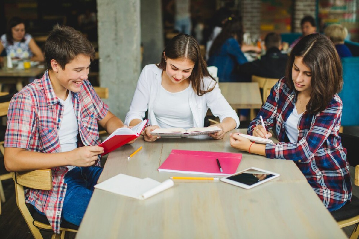 Three students are studying together at a table, surrounded by books, notebooks, and stationery in a lively café setting.