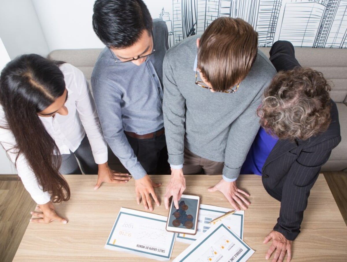 Four professionals collaborate over a table, analyzing data on a tablet and discussing charts and documents in a modern office setting.
