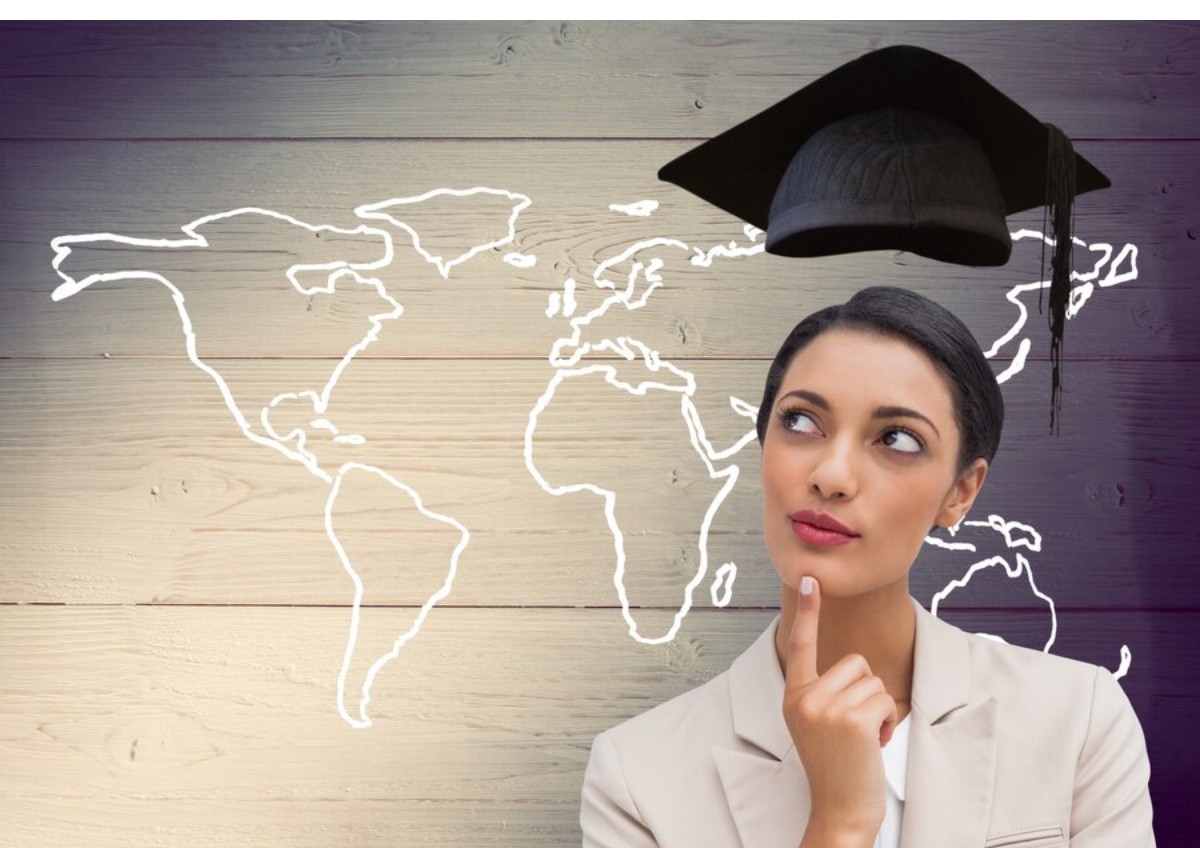 A person in a suit poses thoughtfully with a graduation cap above them, in front of a white outline map on a wooden background.