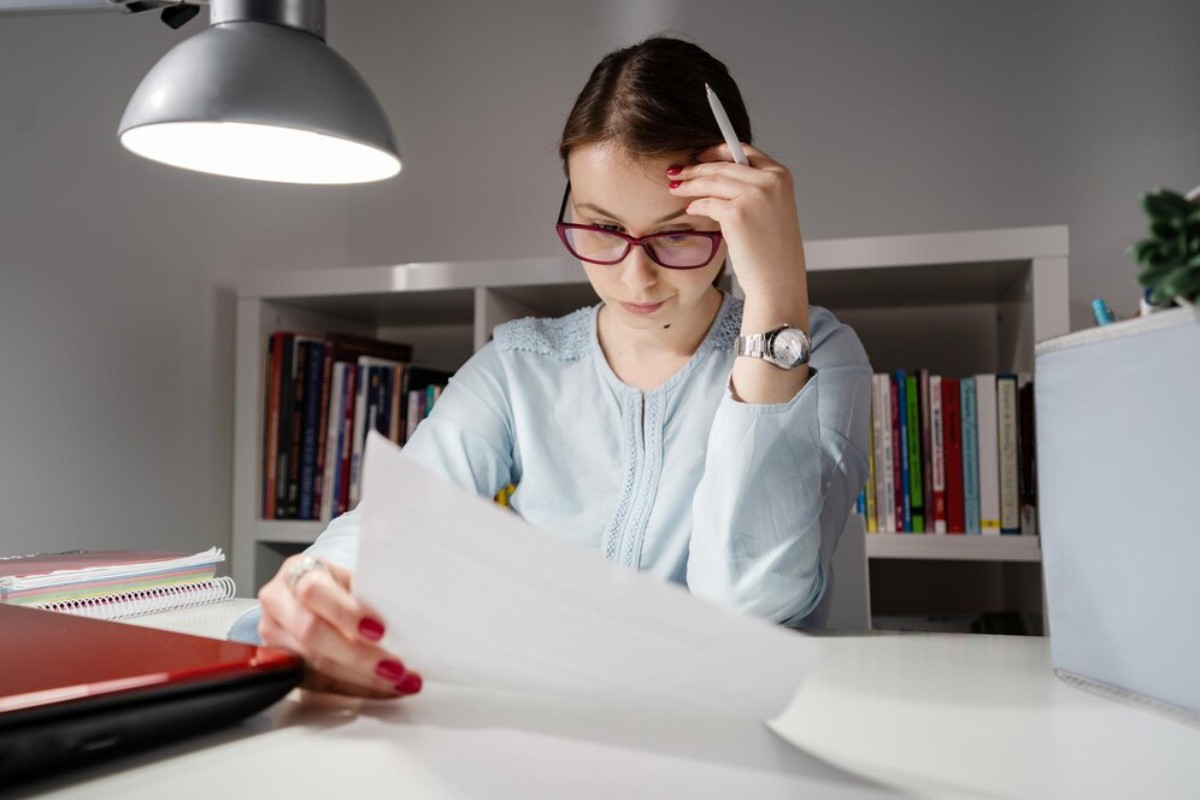 A woman examines a paper while sitting at a desk with a laptop and bookshelves in the background, illuminated by a desk lamp.