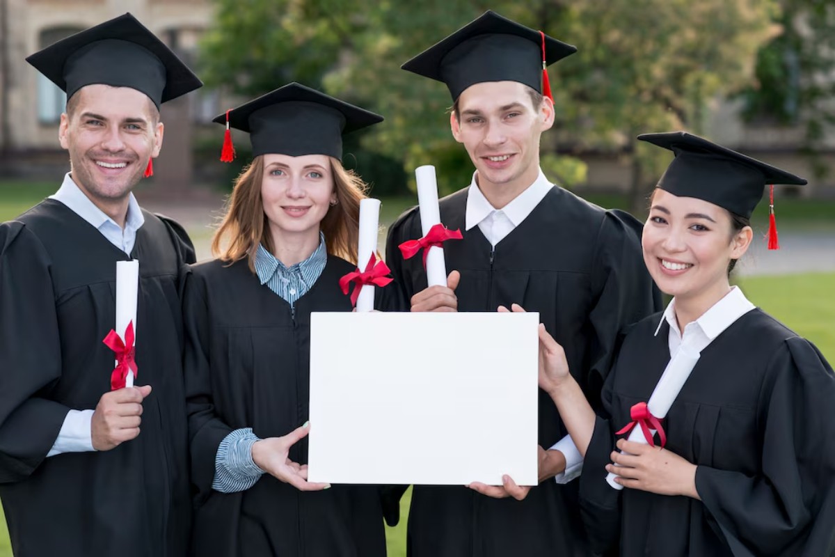 A group of five graduates wearing black caps and gowns proudly hold diplomas and a blank sign in a green outdoor setting.