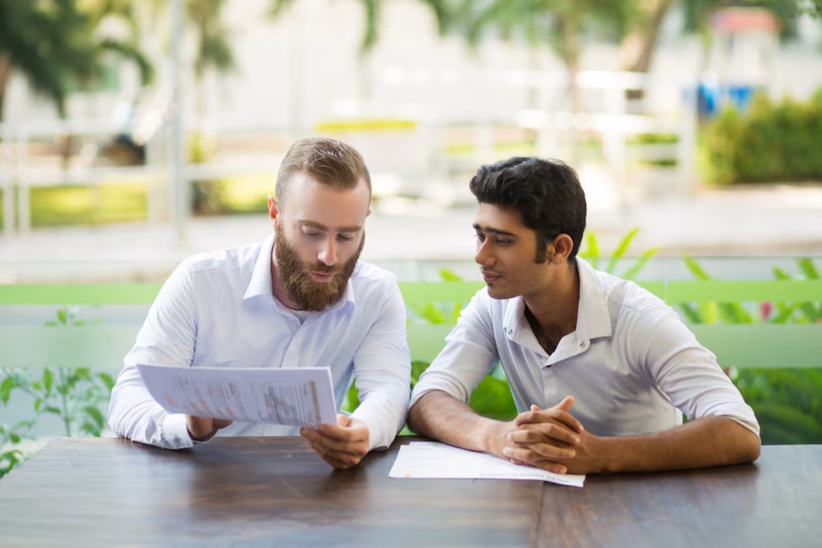 Two men sit at a wooden table outdoors, reviewing documents amidst a lush green background.