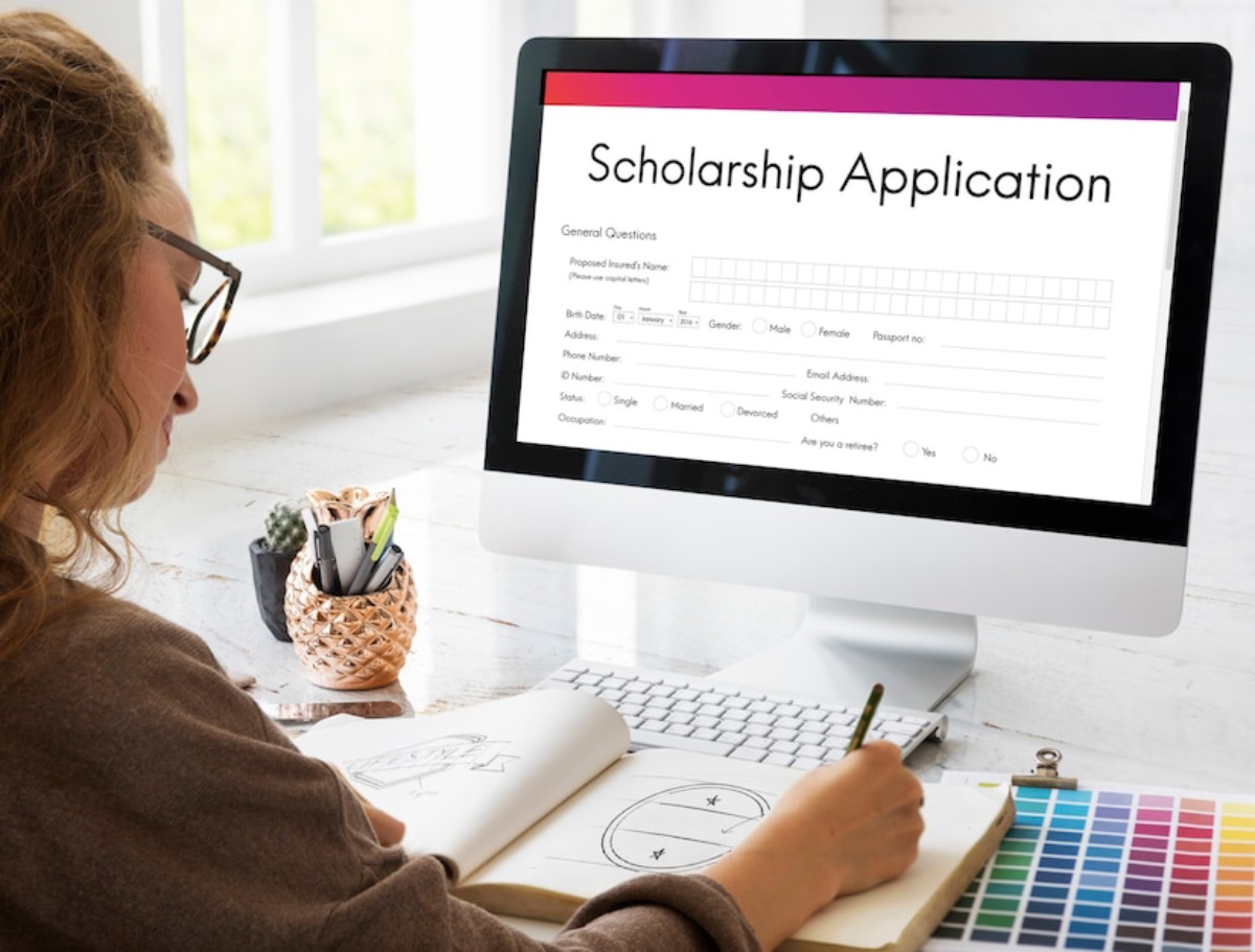 A person fills out a scholarship application on a computer while sketching in a notebook, surrounded by colorful art supplies.