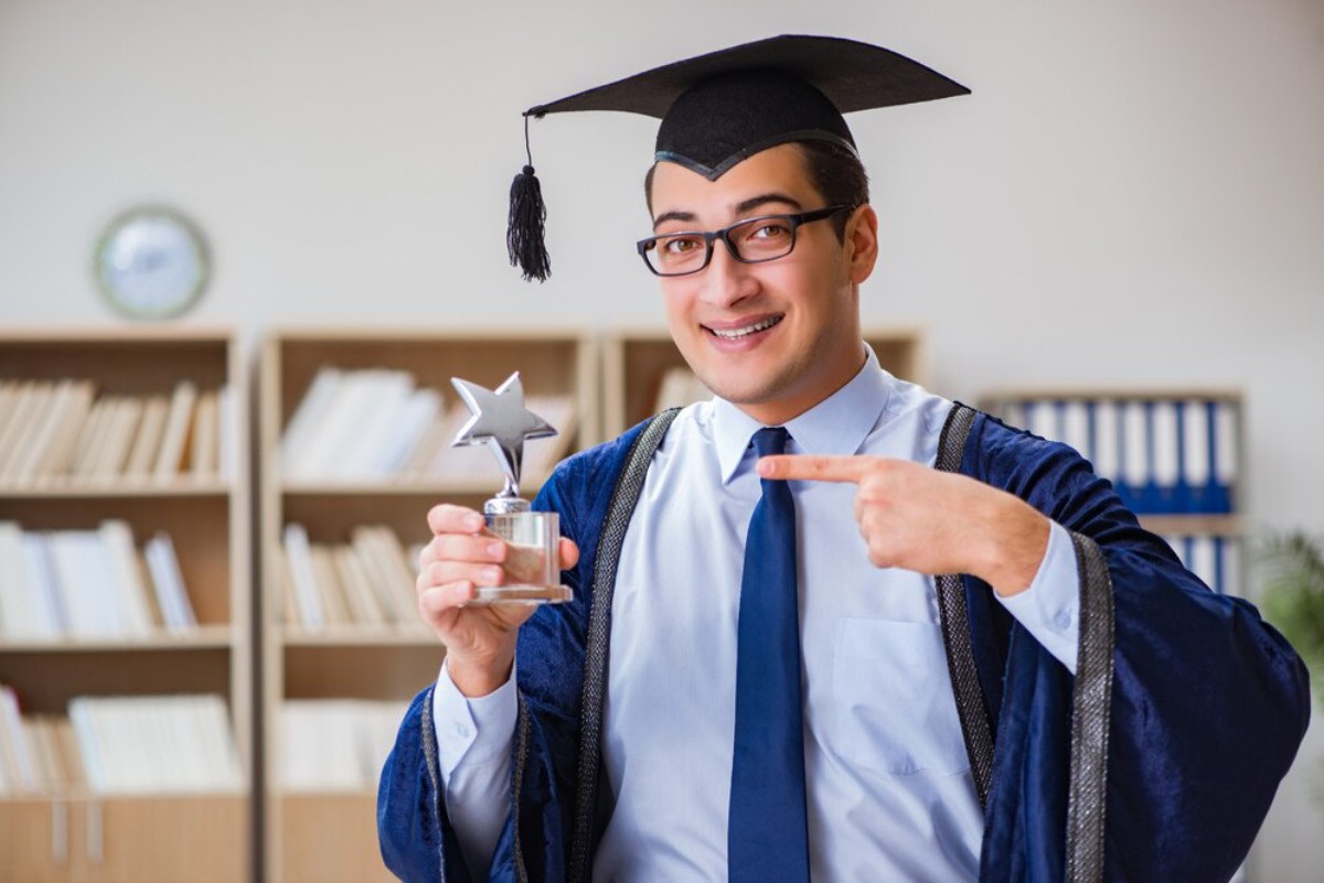 A graduate in a black cap and blue gown holds a trophy with a star, celebrating a significant achievement in a library setting.