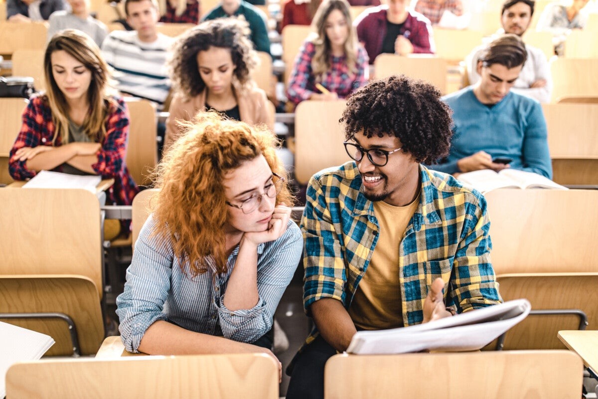 Two students sit closely in a classroom filled with others, engaged in conversation and surrounded by wooden desks and notebooks.