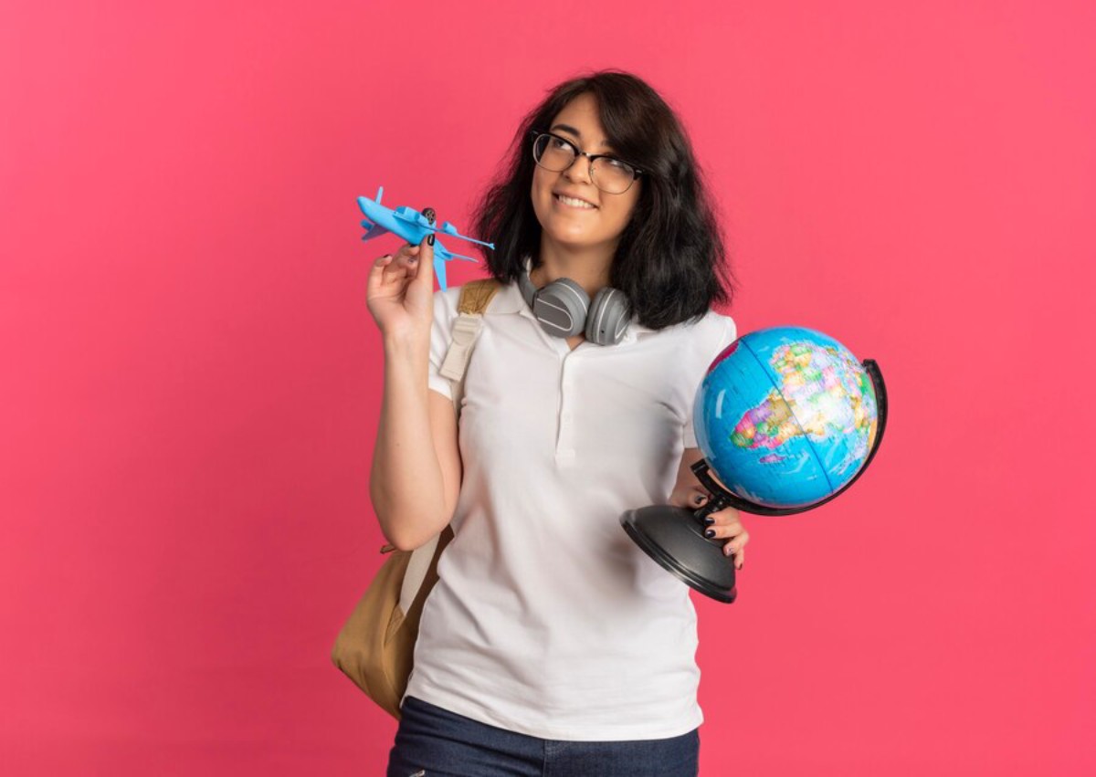 A person holds a blue toy airplane in one hand and a colorful globe in the other, wearing headphones against a pink background.