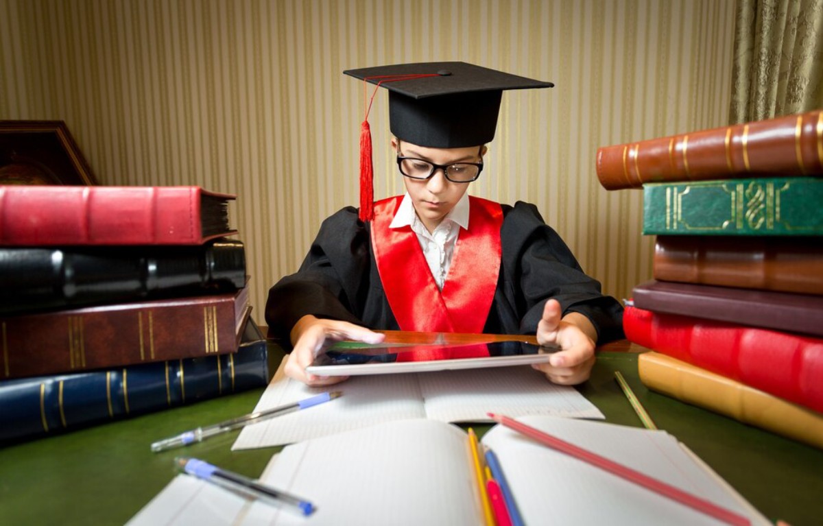 A graduate in a black cap and gown sits at a desk surrounded by books, studying with a tablet and notebooks.