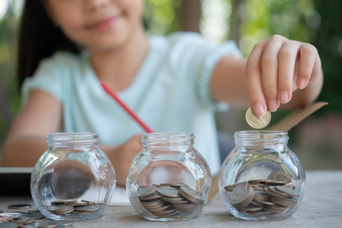 A girl placing a coin into one of three glass jars filled with coins, smiling as she saves money outdoors.