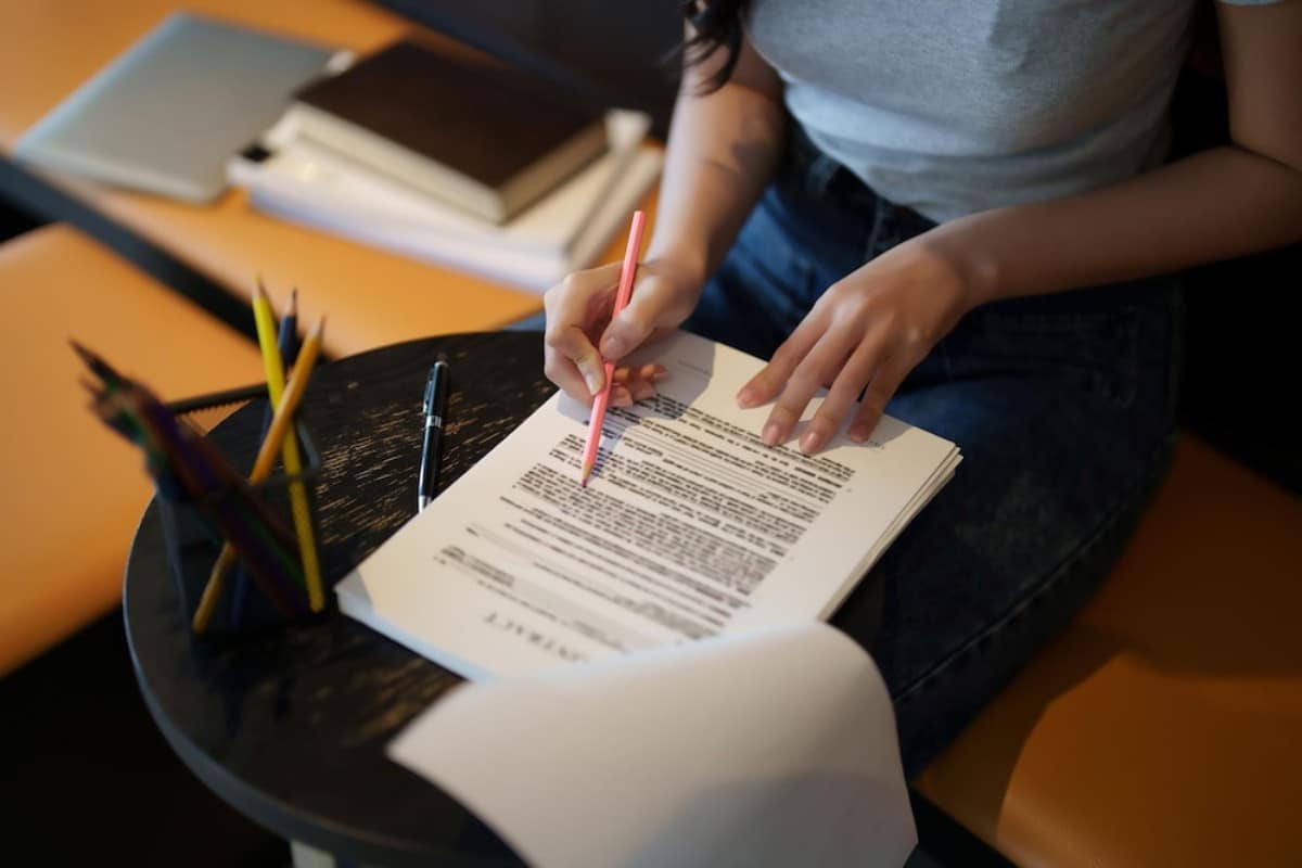A person writing notes in pink pen on a document, surrounded by books and colorful pencils on a small table.