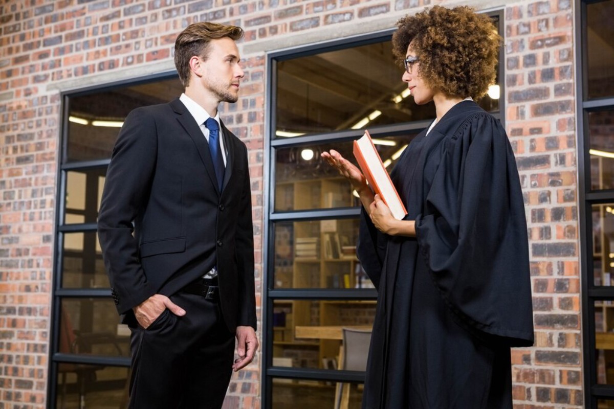 A judge in a black robe holds a book while speaking to a man in a suit, set against a brick wall and large windows.