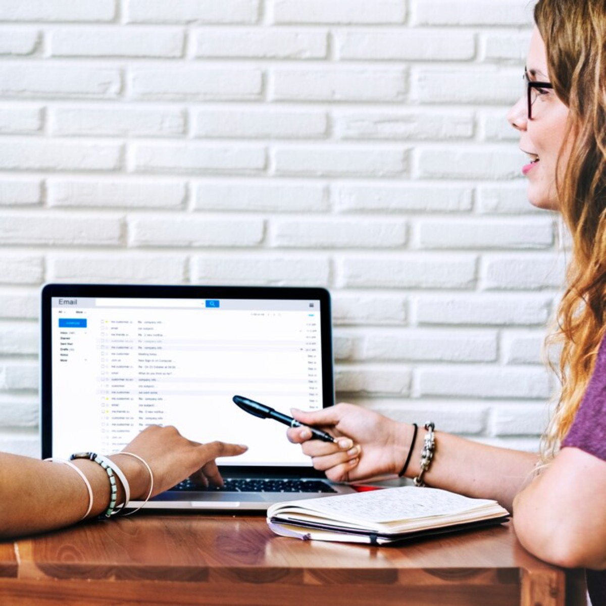 Two people discussing over a laptop displaying an email interface, with a notebook and pen on a wooden table.