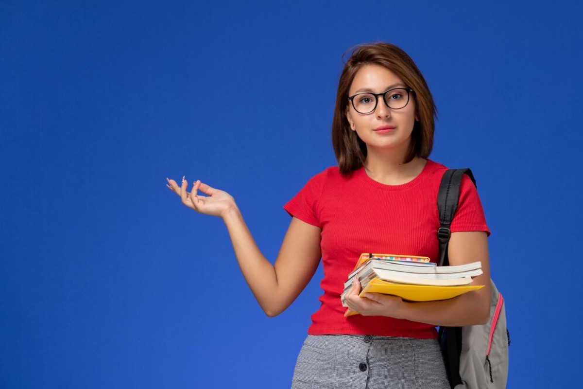 A student in a red shirt holds stacked books and gestures with her arm against a bright blue background.