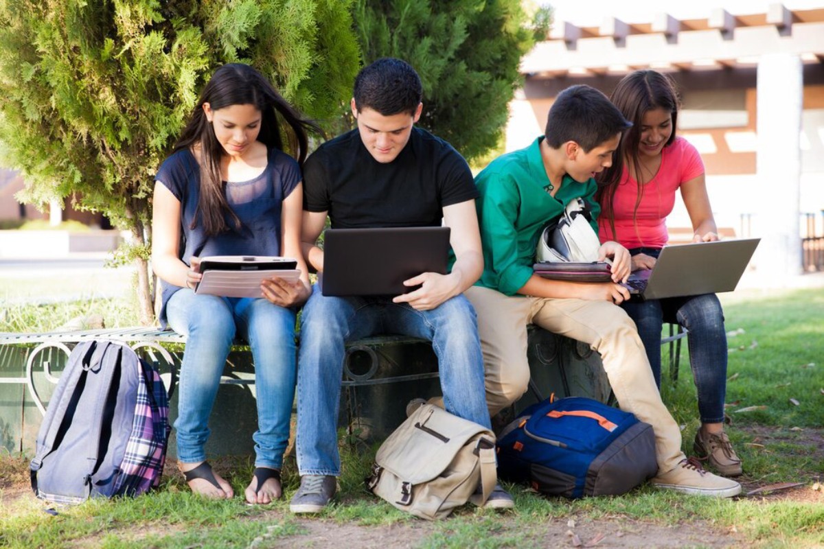 Four young adults sit on a park bench, focused on various electronic devices, surrounded by green trees and backpacks.