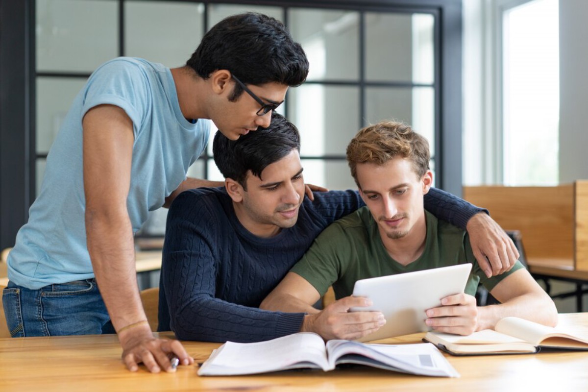 Three students collaborate around a tablet, surrounded by books, in a bright, modern classroom setting.
