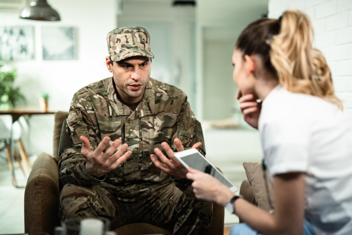 A soldier in camouflage talks passionately while seated, as a woman in scrubs listens intently, holding a tablet.