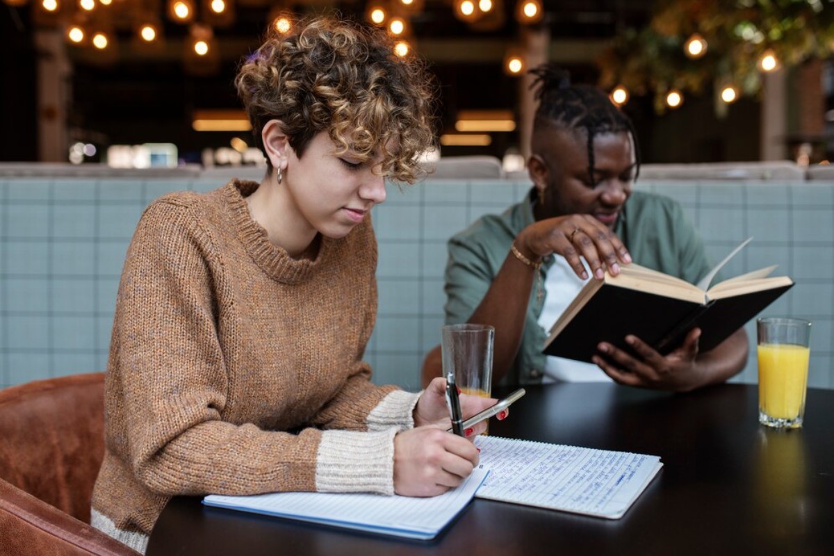 A person with curly hair writes notes at a table, while another reads a book. Drinks sit nearby in a cozy, well-lit café setting.