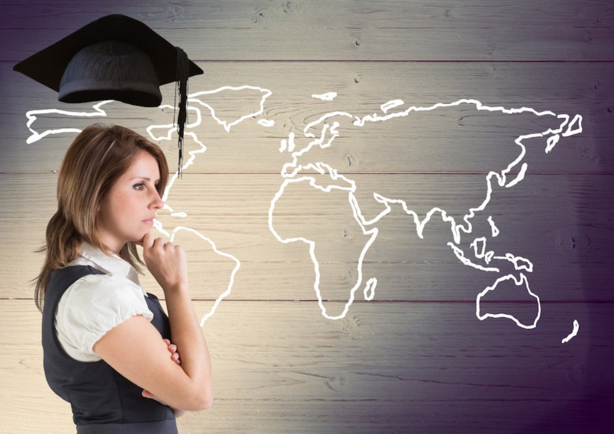 A person in formal attire stands thoughtfully with a graduation cap, in front of a sketched world map on a wooden background.