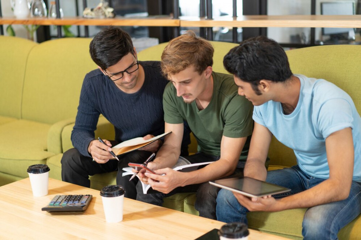 Three young adults engaged in discussion and analysis while seated on a green couch with coffee cups and a calculator on the table.
