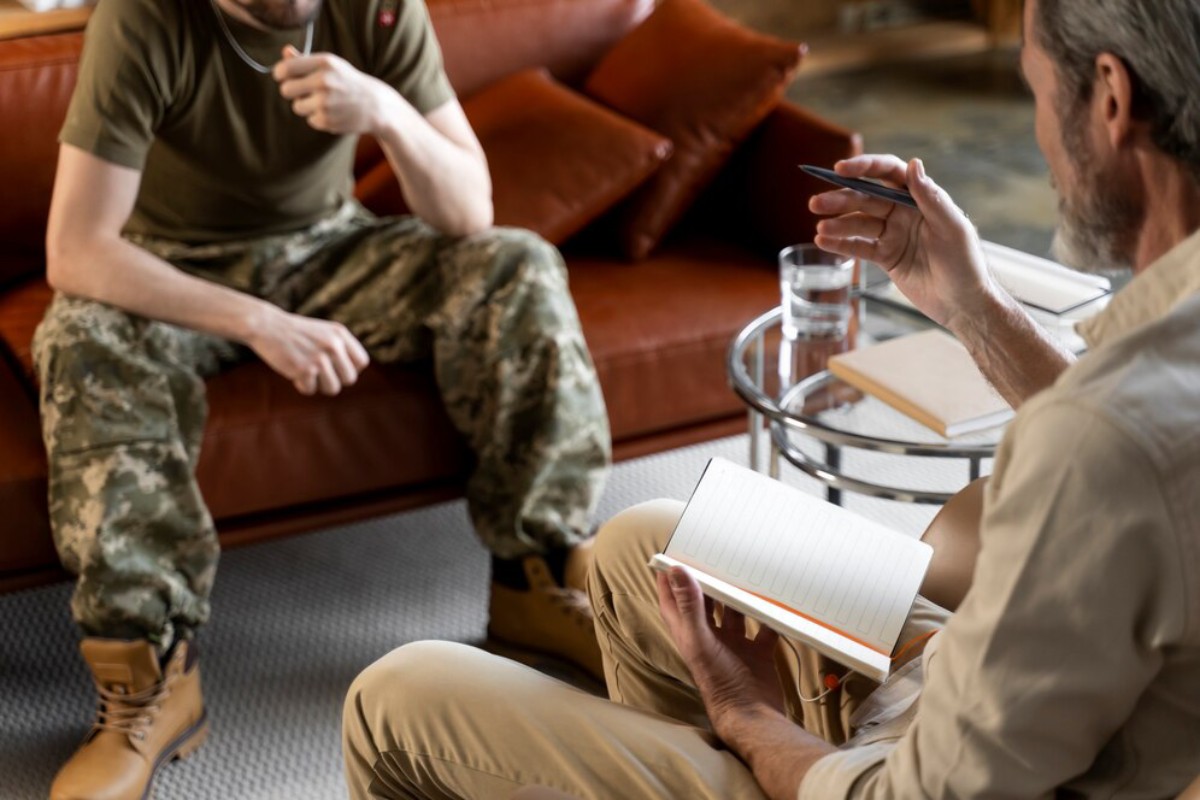 A seated soldier in camouflage speaks thoughtfully with a person in a light shirt, who is taking notes in a notebook.