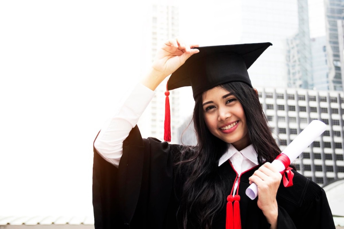A graduate in a black gown and cap, holding a diploma and adjusting their cap, set against a city skyline backdrop.