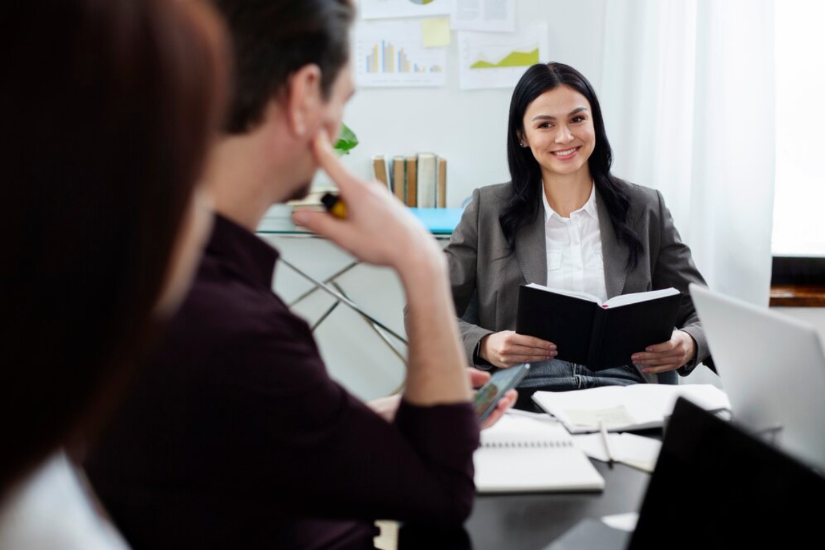 A business meeting with a woman in a suit reading from a book, while two people listen attentively at a conference table.